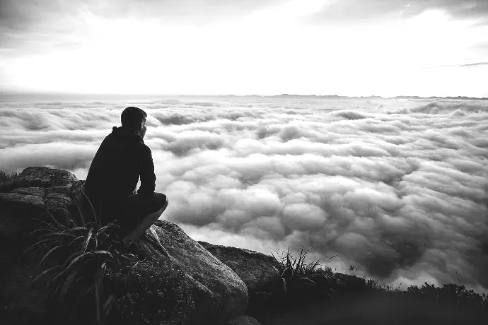 man sitting on gray rock while staring at white clouds